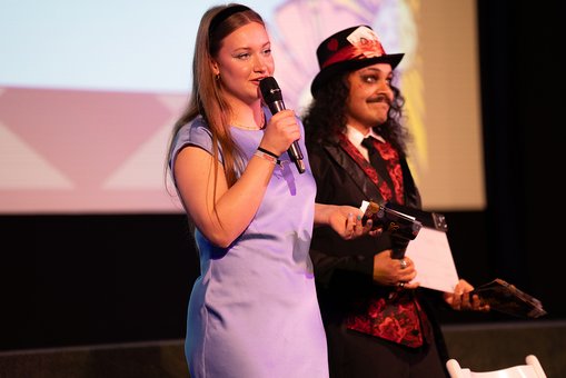 Moderatorin Lilly trägt ein blaues Kleid und hält ein Mikrofon in der Hand, ihr Kollege Joel steht verkleidet als Tonmacher neben ihr