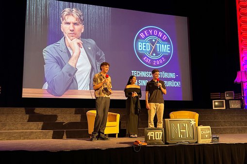 Drei junge Menschen mit Mikrofonen stehen auf einer Bühne vor einer Leinwand und sprechen zum Publikum.