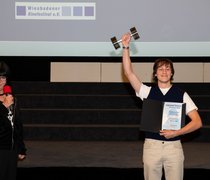 HSRM-Student Moritz Pähler steht während der Preisverleihung des exground filmfest 2024 auf der Bühne und reckt seinen Preis in die Höhe.