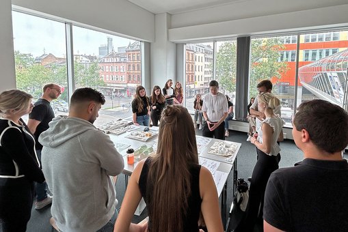Studierende und Lehrende der Hochschule RheinMain stehen um einen Tisch herum und betrachten Architekturmodelle. Im Hintergrund ist durch die Glasfronten die Wiesbadner Innenstadt zu sehen.