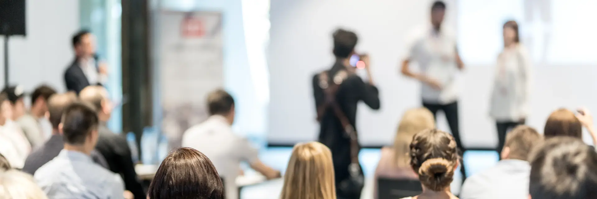 Preisverleihung Symbolbild Leute sitzen in einem großen Raum, auf der Bühne wird ein Foto von Preisträger:in plus Übergabe gemacht
