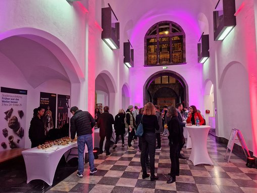 Das Foyer des Museums Wiesbaden erstrahlt in rotem und pinken Licht. An Stehtischen stehen die Besucher:innen mit Getränken und Snacks. 