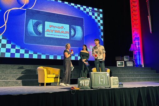 Drei Personen mit Mikrofon stehen auf einer Bühne vor einer Leinwand und sprechen zum Publikum.