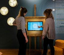 Zwei Frauen stehen im Museum Wiesbaden vor einem Bildschirm und entwerfen mit Touch-Gesten einen Ornament im Jugendstil